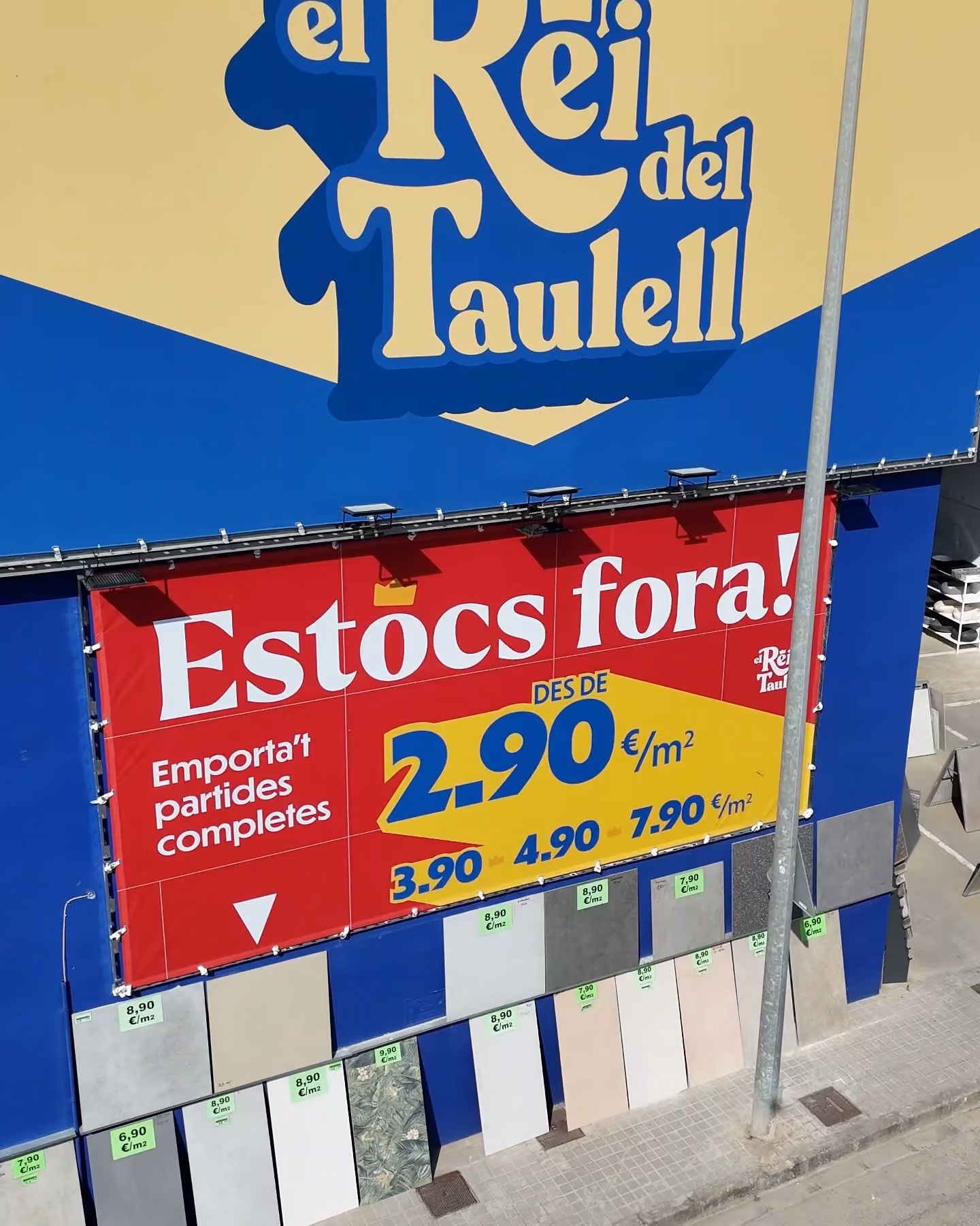 Rótulo de El Rei del Taulell en azul y amarillo, con un cartel rojo de liquidación de estocs a 2,90 euros el metro cuadrado.
